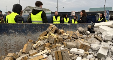 Billedet viser en container med byggeaffald og nogle elever iført gule veste, der modtager undervisning på genbrugspladsen.