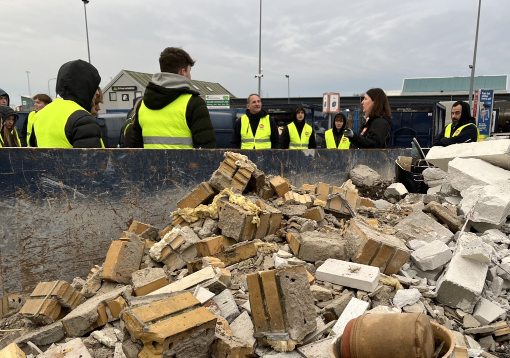 Billedet viser en container med byggeaffald og nogle elever iført gule veste, der modtager undervisning på genbrugspladsen.